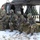 Military personnel in full combat gear are conducting a helicopter insertion/extraction exercise from what appears to be a UH-60 Black Hawk helicopter on a snowy field. Several soldiers wearing mixed camouflage patterns, including DPM (Disruptive Pattern Material) and MultiCam/OCP, are in tactical crouching positions with rifles at the ready. The scene suggests a multinational military training exercise given the variety of uniform patterns visible.