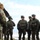 A group of military personnel in full combat gear are gathered in a briefing or pre-mission huddle, likely near a military aircraft given the partial structure visible overhead. One soldier with a visible US flag patch on their left shoulder is facing the group, appearing to address them. The setting appears to be a cold, snowy or frost-covered open terrain.