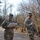 Two U.S. Army soldiers in OCP (Operational Camouflage Pattern) uniforms walk along a gravel path in a wooded area. The soldier on the left is writing on a clipboard while walking, suggesting a field assessment or inspection activity. A third soldier is visible in the background on the same path.