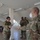 Four U.S. Army soldiers in OCP (Operational Camouflage Pattern) uniforms are gathered inside a building, with two in the foreground exchanging or reviewing documents. One soldier on the right is wearing a white N95-style face mask, suggesting this was taken during the COVID-19 pandemic period. A partially visible sign or checklist is posted on the left side of the frame.