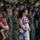 A military ceremony featuring U.S. Marines in MARPAT woodland camouflage uniforms standing in formation alongside civilian family members. The gathering appears to be a formal event such as a change of command, promotion, or unit ceremony, evidenced by the organized formation and presence of families. A woman in a floral dress holds a lei of purple and white orchids, suggesting a Hawaiian or Pacific location.