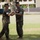 Two U.S. Marines in MARPAT camouflage uniforms are engaged in a formal ceremony on a grassy outdoor area, appearing to exchange or present a ceremonial sword. The interaction suggests a change of command, promotion, or award ceremony typical of military tradition. The setting appears to be a military installation, likely in a tropical or subtropical region based on the vegetation and architecture.