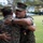 Two U.S. Marines in MARPAT camouflage uniforms are embracing in what appears to be an outdoor military ceremony. The Marine facing the camera is wearing a gold medal with a red ribbon, suggesting a recent award or decoration presentation. Several other Marines are visible in the background observing the moment.