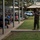 A U.S. Marine in woodland MARPAT camouflage uniform is addressing a mixed audience of military personnel and civilians seated in bleachers under a covered pavilion. The setting appears to be an outdoor military ceremony or briefing on what looks like a military installation, likely in a tropical or warm climate based on the vegetation and building architecture. The speaker is gesturing with both hands raised, suggesting active engagement with the audience.
