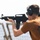 A young man is aiming a black semi-automatic rifle, consistent with an M4/AR-15 style carbine, at a shooting range. He is wearing hearing protection earmuffs and safety glasses, indicating proper range safety protocols. The setting appears to be an outdoor range, possibly on a military vessel or elevated platform based on the background netting.