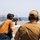 A firearms qualification or training exercise is taking place on the deck of a naval vessel at sea. A person in an orange t-shirt is aiming an M16/M4-style rifle toward paper targets suspended in the distance, while an observer in a tan t-shirt and cap monitors from behind. All visible participants are wearing hearing protection ear muffs.