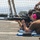 Multiple individuals are in a prone shooting position on a firing range, aiming rifles downrange. The foreground subject is a woman wearing pink ear protection, safety glasses, and a tan/brown shirt, holding what appears to be an M16/AR-15 style rifle. Blue shooting mats are visible beneath the shooters, and a range safety officer or instructor is partially visible at the right edge.