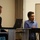 Two individuals are seated or standing at a table/podium in what appears to be a presentation or seminar setting. The person on the right, wearing a blue sweater and glasses, appears to be speaking or listening attentively, while the person on the left in a dark blazer looks downward. A laptop and a whiteboard are visible in the background.
