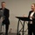 Two professionals are presenting in what appears to be a conference or meeting room setting. A woman in a dark blazer stands at a black podium gesturing with her hands while speaking, while a man in a dark suit sits behind an HP laptop at a separate stand. The setting suggests a formal presentation or panel discussion.