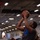 The Harry S. Truman Carrier Strike Group is on a scheduled deployment in the U.S. Sixth Fleet area of operations in support of naval operations to maintain maritime stability and security. A basketball game is being played inside what appears to be an aircraft carrier hangar bay, with military aircraft visible in the background. One player wearing NBA All-Star shorts is going up for a shot near the basket while a defender attempts to contest. A crowd of spectators watches from the sidelines.