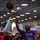 The Harry S. Truman Carrier Strike Group is on a scheduled deployment in the U.S. Sixth Fleet area of operations in support of naval operations to maintain maritime stability and security. A basketball game is being played inside what appears to be a naval aircraft carrier hangar bay, with military aircraft visible in the background. A player in a purple shirt is shooting the ball toward the basket with 6 seconds remaining on the clock. The scoreboard shows the home team leading 60 to 2, and a crowd of spectators lines the background.