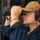 The Harry S. Truman Carrier Strike Group is on a scheduled deployment in the U.S. Sixth Fleet area of operations in support of naval operations to maintain maritime stability and security. A young military service member is looking through large binoculars while wearing a tan/khaki cap with patches and communication headphones. The individual is dressed in a dark navy blue uniform and wears glasses and a gold chain bracelet. The setting appears to be aboard a naval vessel, likely on a bridge or observation deck.