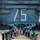 The Harry S. Truman Carrier Strike Group is on a scheduled deployment in the U.S. Sixth Fleet area of operations in support of naval operations to maintain maritime stability and security. A large group of personnel are assembled on the flight deck of a U.S. Navy aircraft carrier numbered '75', holding up blue cards or papers above their heads. They are arranged in a formation that appears to spell out or form a shape visible from above. The ship's island superstructure is visible in the background with the hull number '75' and a safety warning reading 'BEWARE OF JET BLAST PROPELLERS AND ROTORS'.
