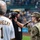 A female U.S. Air Force service member in OCP (Operational Camouflage Pattern) uniform is speaking into a microphone at what appears to be a Pittsburgh Pirates baseball game. A person wearing a Pirates jersey with the name 'GRUSHECKY' and number 1 stands in the foreground, while a videographer captures the moment. The stadium is filled with spectators in the background.