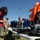 Military personnel in camouflage uniforms are conducting an equipment demonstration or display on a grass field, likely at a school or community outreach event. A civilian student with a backpack observes as soldiers explain various pieces of equipment laid out on folding tables. An orange hazmat/chemical protective suit is prominently displayed on a rack attached to a dark blue military vehicle.