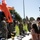 Military personnel in camouflage uniforms are conducting an outdoor demonstration or educational display for a group of civilians. A prominent orange hazmat/chemical protective suit is displayed on the back of a dark emergency response vehicle. Various detection and response equipment is laid out on a table in the foreground.