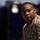 A U.S. Army soldier wearing Operational Camouflage Pattern (OCP) uniform is photographed in a close-up portrait style. The soldier's uniform displays a 'CBRN' (Chemical, Biological, Radiological, and Nuclear) tab patch and a U.S. Army eagle emblem on the left shoulder. The background is heavily blurred with blue and dark tones, suggesting an outdoor military or industrial setting.