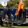 Military personnel are conducting what appears to be a public demonstration or outreach event on a grassy field. Two soldiers in camouflage uniforms are presenting equipment at a folding table to two civilians. A prominent orange hazmat/CBRN protective suit is displayed on top of a blue emergency response vehicle, alongside a satellite communication dish on a second vehicle.