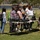 A military service member in camouflage uniform is presenting equipment to a group of civilians, likely students, at an outdoor demonstration on a grass field. The group is gathered around folding tables displaying various pieces of military or technical equipment. The setting appears to be an educational outreach or recruitment event.
