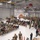 A large military ceremony or formation is taking place inside a spacious hangar facility. Hundreds of uniformed military personnel and civilian guests are gathered, with some seated in folding chairs and others standing, facing a stage with a podium where a speaker appears to be addressing the crowd. Multiple military flags are visible on the stage, suggesting an official ceremony such as a deployment, redeployment, or unit activation event.