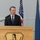 A man in a dark suit and blue striped tie is speaking at a wooden podium equipped with a microphone. Behind him are two flags: a United States flag and what appears to be a blue organizational or state flag. The podium features an emblem depicting a profile of a figure, possibly a Native American or classical figure, within a shield shape.