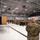 A military ceremony is taking place inside a large hangar facility, with a uniformed soldier speaking at a wooden podium on a raised stage. A large audience of military personnel and civilians is seated in folding chairs facing the speaker, while another soldier stands in the foreground with their back to the camera. The setting appears to be a formal military event such as a change of command, activation, or unit ceremony.