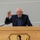 An elderly man with white hair and glasses is speaking at a wooden podium equipped with a microphone. The podium bears the emblem of 'The Green Mountain Boys,' featuring a colonial-era figure holding a musket. The speaker is gesturing with his right hand raised, appearing to be mid-speech.