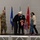 A military ceremony is taking place on a stage, where two civilians in formal attire appear to be presenting a Blue Star Service Banner to a young girl, with her family present. A uniformed military service member stands at a podium bearing the 'Green Mountain Boys' insignia. The setting includes multiple flags including the US flag, Air Force flag, and a general officer flag with two stars.