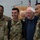 An elderly civilian man in a blue sweater and glasses poses for a photo with three U.S. Air Force service members in OCP (Operational Camouflage Pattern) uniforms. The group is smiling and appears to be at an indoor military facility or event space, with a podium and flags visible in the background. The atmosphere is friendly and informal.