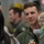 A young male military aviator in an olive green flight suit is smiling and holding a small infant wearing a knit beanie hat. The setting appears to be a hangar or military facility, with other personnel in flight suits visible in the blurred background. The scene suggests a homecoming or family reunion event.