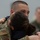 A U.S. Army soldier in MultiCam/OCP camouflage uniform is embracing a person with dark hair, in what appears to be an emotional reunion or farewell moment. The soldier wears a U.S. flag patch and unit patch on the left sleeve. A second uniformed individual is visible but blurred in the background.