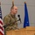 A military officer in OCP (Operational Camouflage Pattern) uniform is speaking at a wooden podium with a microphone. The setting appears to be an official military ceremony or briefing, with multiple flags visible in the background including the United States flag and what appears to be Air Force and unit flags. The individual has a U.S. flag patch visible on the right sleeve of their uniform.