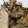 Two military personnel in MARPAT (Marine pattern) camouflage uniforms are positioned behind a wooden barrier in what appears to be a training exercise. The foreground figure is kneeling and holding an M16/M4-style rifle while wearing full tactical gear including a ballistic helmet, plate carrier, and eye protection. The setting appears to be an outdoor training range with open grass fields and tree lines in the background.