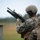 Two U.S. Marines in MARPAT digital camouflage uniforms are at what appears to be a firing range or training exercise. The foreground Marine is holding an M16/M4-style rifle with an M203 grenade launcher attachment and is loading or handling a 40mm grenade round. A second Marine, identified by the name tape 'WHITEHEAD,' observes from behind.