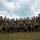 A large group of military personnel, approximately 25-30 individuals, pose for a formal unit group photograph outdoors. They are wearing digital camouflage uniforms consistent with U.S. Marine Corps MARPAT pattern, tactical vests, and combat helmets. The group is arranged in multiple rows with some seated in front and others standing behind, several holding rifles raised.