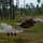 The image shows a military training range with a stationary metal tank silhouette target and several human-shaped steel targets positioned across an open field. Smoke plumes are visible near the targets, indicating recent weapons fire or explosive simulation. The setting appears to be a live-fire training area surrounded by sparse pine forest.