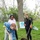 Three young women are participating in a tree-planting event, digging a hole to plant a balled-and-burlapped tree sapling. A 'Tree City USA - Arbor Day Foundation' sign is visible in the background, indicating this is an organized community planting event. The scene takes place in a grassy park-like setting with existing trees and early spring foliage.