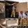 A group of firefighters in turnout gear are conducting what appears to be a training exercise involving a modified shipping container or trailer unit in an arid, desert-like environment. One firefighter is inside the container manipulating a large wooden panel or door, while three others stand outside observing or preparing to assist. The setting, equipment wear patterns, and training structure suggest a live fire or forcible entry drill.