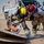 Two firefighters or rescue personnel in turnout gear are using an orange rotary rescue saw (cut-off saw) to cut through wooden planks, likely during a training exercise in an arid desert environment. A third person in blue uniform observes in the background near a large tan/desert-colored fire or rescue truck. The scene appears to be a technical rescue or extrication training drill.