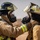 Two firefighters in full personal protective equipment face each other, with one adjusting or checking the breathing apparatus mask of the other. Both wear turnout gear with high-visibility yellow reflective striping, SCBA (Self-Contained Breathing Apparatus) tanks, and full-face respirator masks. The setting appears to be an arid, outdoor environment, suggesting a training exercise or operational deployment.