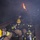 A group of firefighters in full protective gear are crouched low inside a smoke-filled structure with active flames visible near the upper ceiling area. The scene appears to be either a live fire training exercise or an active structural fire response. The firefighters are working in close proximity to one another in a low-visibility, high-hazard environment.