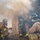 Three firefighters in full personal protective equipment are conducting what appears to be a firefighting operation or training exercise near a smoke-filled doorway or structure. They are wearing MSA brand self-contained breathing apparatus (SCBA) and standard turnout gear with high-visibility reflective striping. Heavy smoke is billowing from the structure, and a fire hose is visible on the ground.