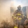 Three firefighters in full personal protective equipment (PPE) are emerging from a smoke-filled environment, likely during a live fire training exercise. The central firefighter is standing and moving forward while two others are crouched low near the ground. Dense white and gray smoke fills the background, partially obscuring a metal structure or container.