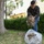 A young man wearing a camouflage jacket and jeans is collecting dead leaves and yard debris into a large clear plastic bag outdoors. He is bending forward, dropping a handful of dry leaves and plant material into the partially filled bag. The setting appears to be a courtyard or garden area near European-style buildings.