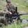 Two U.S. Army soldiers in MultiCam (OCP) camouflage uniforms are working with a crew-served weapon in a field training environment. The soldiers appear to be conducting maintenance, assembly, or familiarization training on what appears to be an M2 .50 caliber heavy machine gun mounted on a tripod. The setting is an outdoor training range with grass and sandy terrain.