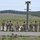 A group of approximately 8-9 military personnel in camouflage uniforms are gathered at what appears to be a firing range or training area. They are standing near a utility pole equipped with cameras and lights, with a wide open field extending into the background. Smoke or fire is visible in the far background, suggesting active range use.