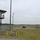 The image shows a military training range facility with a two-story observation/control tower on the left side. A military HMMWV (Humvee) vehicle is positioned in the middle distance, with a group of soldiers gathered nearby, apparently conducting a training exercise. The facility features open flat terrain, chain-link fencing, utility poles, and a tree line in the background under an overcast sky.