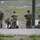 A group of military personnel in camouflage uniforms are conducting what appears to be a weapons training exercise at an outdoor firing range. Several soldiers are positioned near shooting lanes marked with target boards, with some observing and others actively engaged in range activities. Ammunition boxes and linked ammunition are visible on the ground nearby.