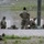 Military personnel in MultiCam/OCP camouflage uniforms are conducting a live-fire range training exercise at what appears to be a military shooting range. Several soldiers are in prone firing positions while others stand or kneel in supervisory or observing roles. Paper silhouette targets labeled 'A' and 'D' are visible downrange, and ammunition cans are present on the ground.