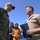 Two U.S. Navy personnel are engaged in conversation outdoors under a clear blue sky. One is wearing Navy Working Uniform (NWU) camouflage with a name tape reading 'STEWART' and a baseball cap bearing small flag patches, while the other is in the tan Service Khaki uniform displaying multiple medal ribbons and holding a dark folder. Two civilians, appearing to be a woman and a young girl, stand in the background observing the exchange.
